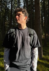 Ung mann iført svart Salomon T-skjorte og ryggsekk står i en solfylt tett skog med høye trær og grønn mose på bakken.