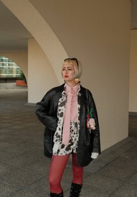 Persoon met een blonde bobstijl, gekleed in een zwart leren jack, roze blouse en strik, koeienprint shorts, rode panty's en zwarte laarzen, die onder een boog staat.
