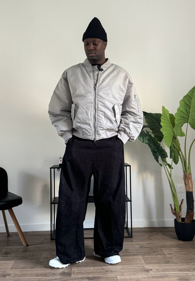 Veste bomber gris clair avec des poignets côtelés, pantalon noir oversize, baskets blanches et un bonnet en tricot noir, se tenant devant un fond minimaliste.