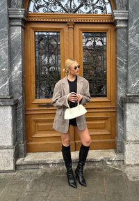 Ruitvormige oversized blazer in lichtbruin, zwarte shorts, kniehoge zwarte laarzen en een crèmekleurige handtas. Een deur met een metalen draadontwerp dient als achtergrond.