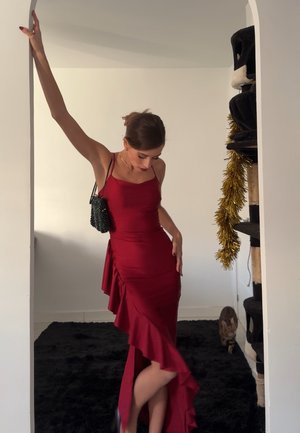 Vestido de satén rojo con tirantes finos, que presenta un dobladillo con volantes y un detalle lateral. Un pequeño bolso negro con cuentas añade contraste. Iluminación suave en interiores.
