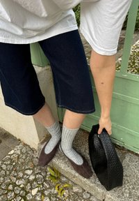 Mørk denim bermuda shorts, grå ribbede sokker, brune flade sko, og en struktureret sort clutch mod en grøn træbaggrund.