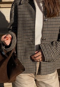 Hnedo-biely kabát s vzorom psieho zubčeka, veľkými klopami a zapínaním na gombíky, skombinovaný s krémovým sveterom a hnedou semišovou kabelkou.