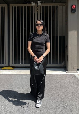 Schwarzes tailliertes Kurzarmhemd, schwarzer langer Rock, schwarze Handtasche, silberner Schmuck, Sonnenbrille und schwarz-weiße Sneakers vor einem zaunbewehrten Hintergrund.