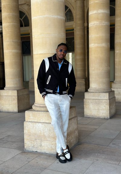 Hombre vestido con chaqueta blanca y negra, pantalones blancos y zapatos mocasines, apoyado contra una columna de piedra en un área con grandes pilares de piedra.