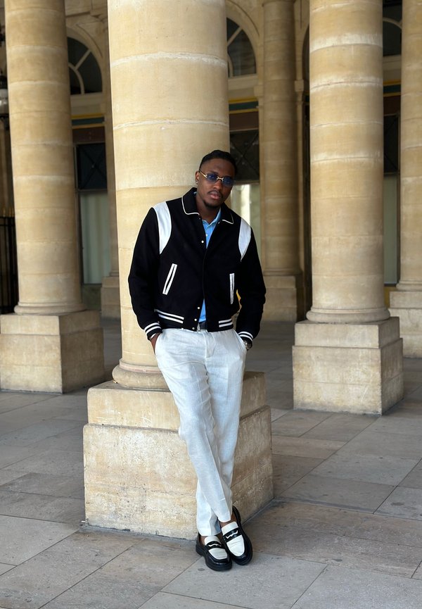 Homme portant une veste noire et blanche, un pantalon blanc et des mocassins, appuyé contre une colonne en pierre dans une zone avec de grandes colonnes en pierre.