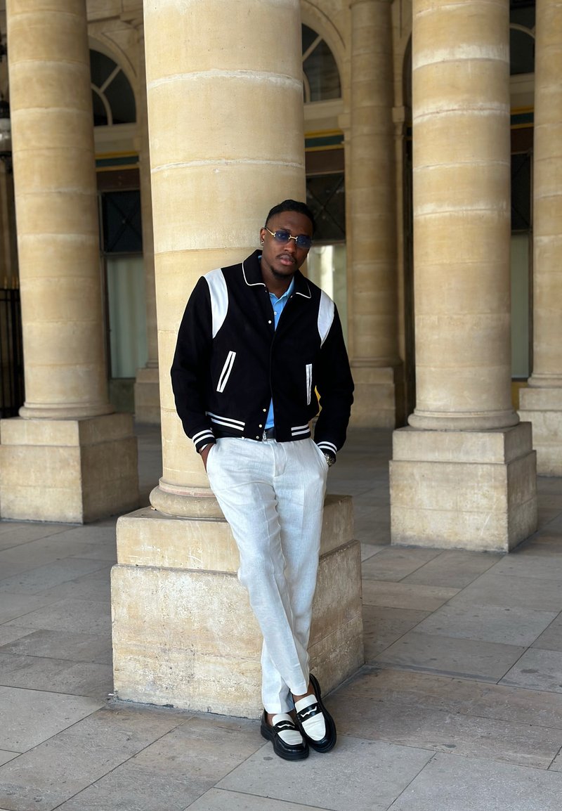 Moški, oblečen v črno-belo jakno, bele hlače in natikače, se naslanja na kamniti steber v prostoru z velikimi kamnitimi stebri.