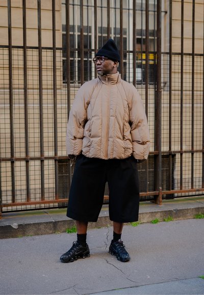 Chaqueta de plumas color canela con un alto cuello, pantalones cortos negros de gran tamaño y zapatos negros con diseño texturizado. Los accesorios incluyen un gorro negro y gafas.