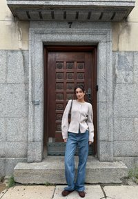 Cardigan in lana beige morbida con orlo arrotondato e bottoni, abbinato a jeans a gamba larga azzurri chiaro e mocassini marroni. Sfondo con porta in pietra.