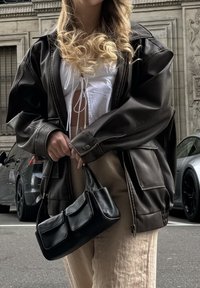 Καφέ δερμάτινο υπερμεγέθες μπουφάν, άσπρη μπλούζα με λεπτομέρειες δέσιμο, μπεζ παντελόνι και μαύρη τσάντα σε στυλ χειρός με δύο μπροστά τσέπες. Αστική ατμόσφαιρα.