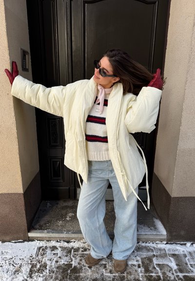Mujer con ropa de invierno, incluyendo chaqueta acolchada blanca, suéter a rayas, guantes rojos, gafas de sol y jeans claros, posando junto a una puerta oscura en la nieve.