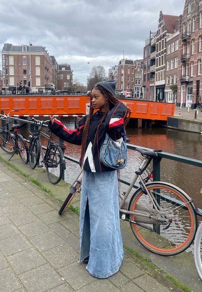 Falda larga de mezclilla, chaqueta negra oversize con acentos rojos y blancos, bolso azul, gorro de punto gris, cabello trenzado. Bicicletas y canal de fondo.