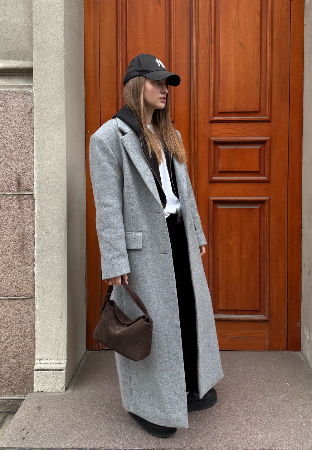 Manteau long en laine mélangée gris, associé à un hoodie noir et une chemise. Porte un sac à main en daim marron foncé. Fond de porte marron.