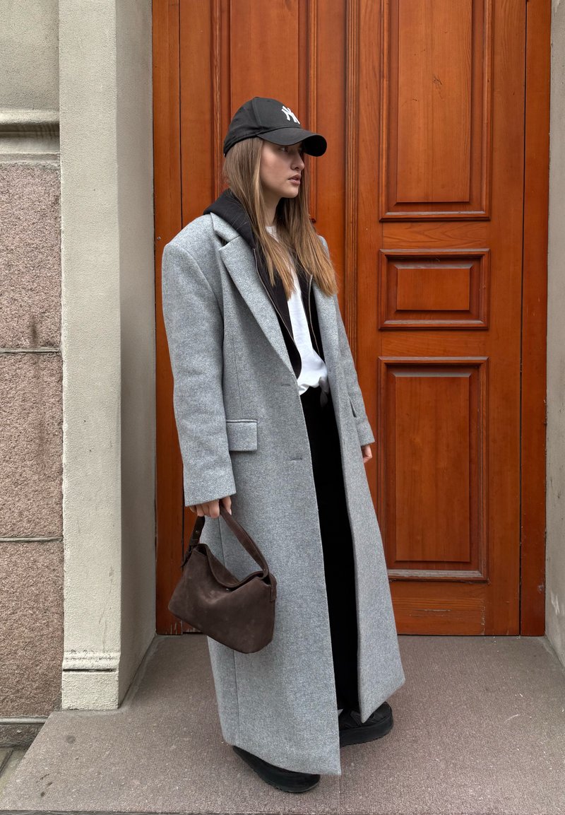 Manteau long en laine mélangée gris, associé à un hoodie noir et une chemise. Porte un sac à main en daim marron foncé. Fond de porte marron.