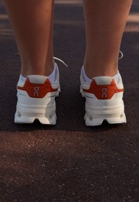 Weiße Sport Schuhe mit orangefarbenen Akzenten, strukturierten Einsätzen und Branding-Details. Das Profilmuster auf der Sohle ist sichtbar und wurde zur Unterstützung entwickelt.