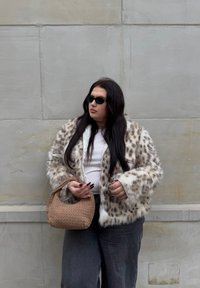 Giacca di pelliccia finta con un motivo maculato in beige e bianco, abbinata a una maglietta bianca e jeans in denim scuri. Tieni una borsa intrecciata marrone.