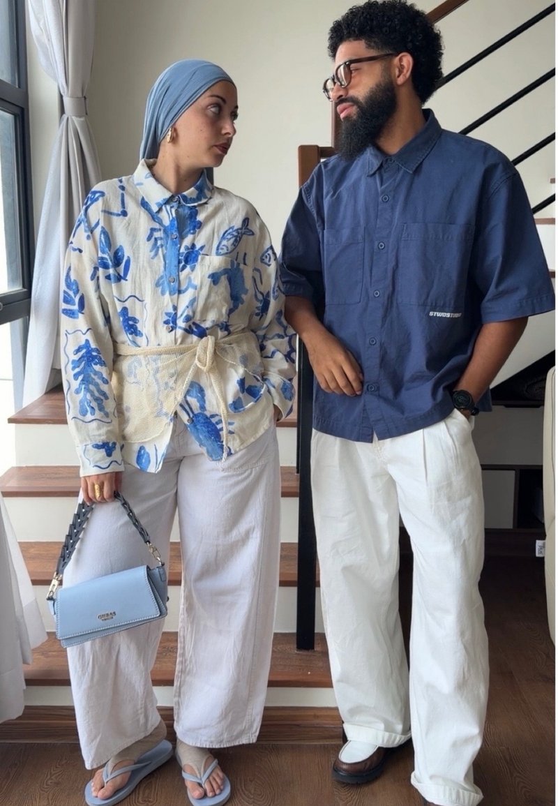 Femme en chemise fleurie et foulard tenant un sac bleu et homme en chemise bleue et pantalon blanc se tiennent face à face à l'intérieur près des escaliers.