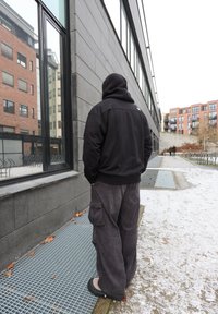 Czarna bluza z kapturem, szare spodnie cargo oraz ciemne obuwie. W tle nowoczesny budynek z dużymi oknami i elementami z cegły.