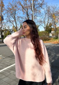 Young woman with long dark hair wearing a pink jumper stands outdoors on a sunny autumn day with bare trees in the background.