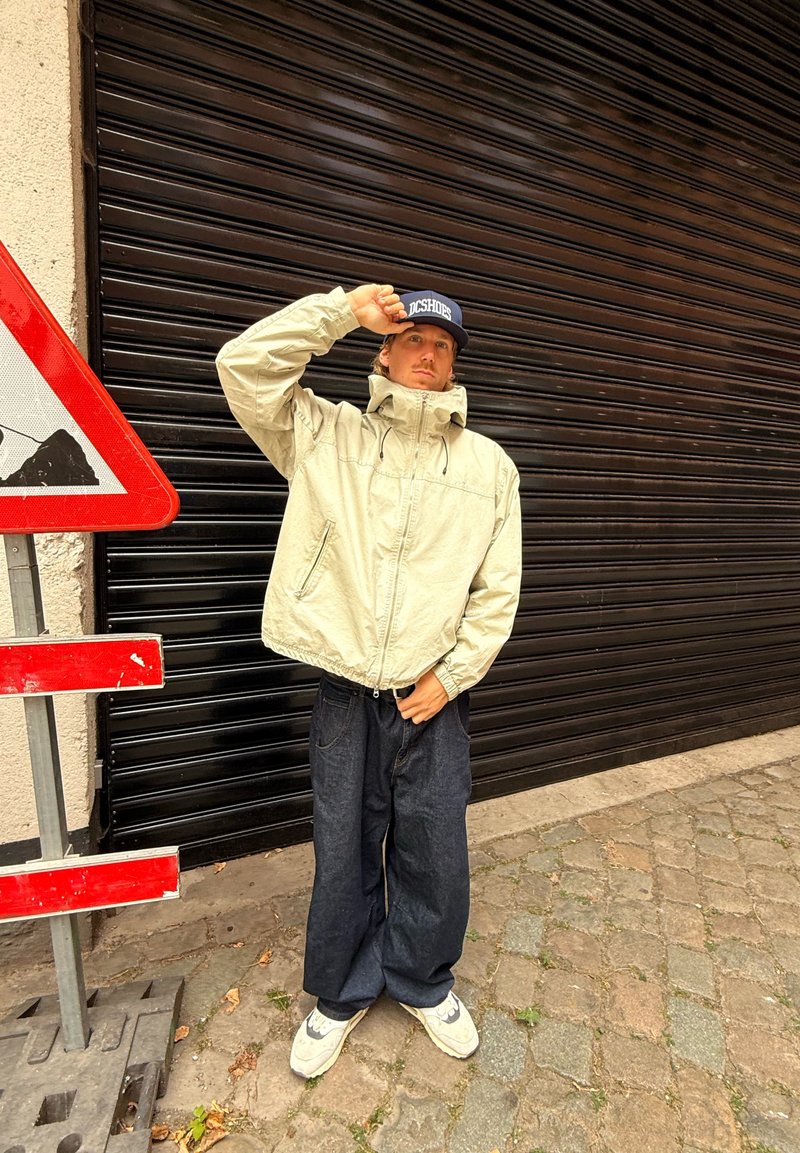 Lys beige zip-up jakke med høj krave, parret med løse mørke denimbukser og hvide sneakers. Kasketten har "DC Shoes" logo.