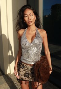 Fiatal nő ezüst flitteres pánt nélküli felsőben és gyöngyökkel díszített virágmintás miniszoknyában, kezében nagy barna szőrme táskával, napsütésben áll kint.