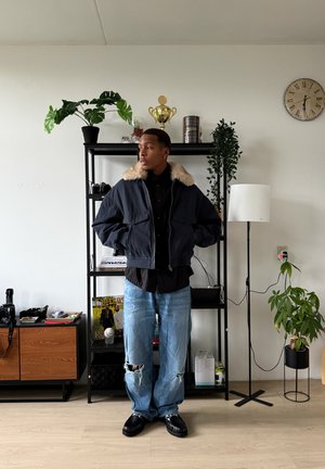 Chaqueta bomber azul marino con cuello de piel sintética, camisa negra abotonada, vaqueros desgastados azul claro y zapatos negros, de pie en una habitación amueblada.