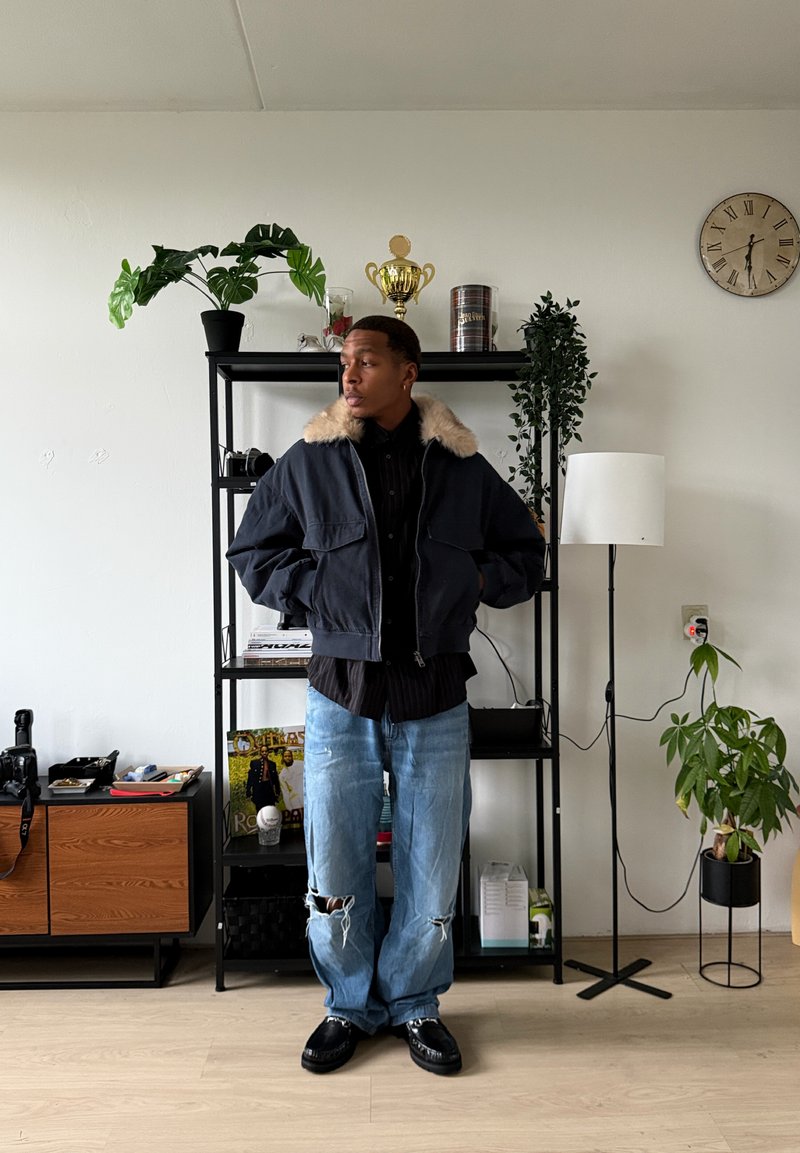 Blouson aviateur bleu marine avec col en fausse fourrure, chemise noire à boutons, jean bleu clair déchiré et chaussures noires, debout dans une pièce meublée.