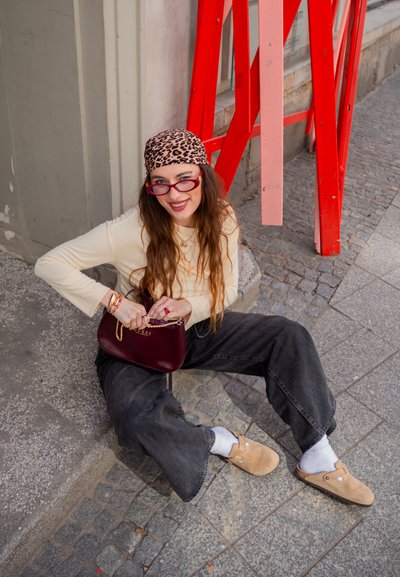 Joven mujer con cabello largo y pañuelo de estampado de leopardo sentada en la acera urbana, sosteniendo un bolso burdeos, llevando gafas, camiseta beige y pantalones negros.