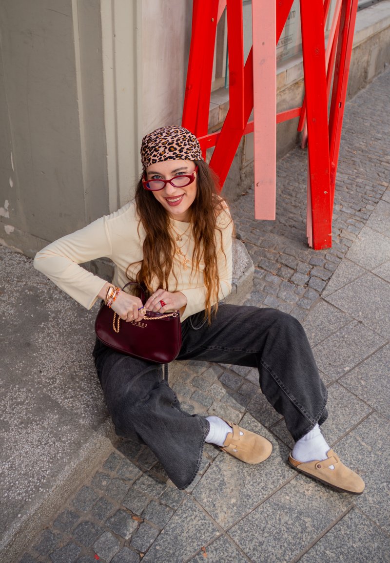 Mladá žena s dlouhými vlasy a šátkem s leopardím vzorem sedí na městském chodníku, drží vínovou kabelku, má brýle, béžový top a černé kalhoty.