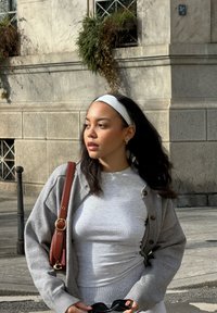 Cardigan gris avec de grands boutons sur un haut gris. Sac à épaule en cuir marron, bandeau blanc et lunettes de soleil noires à la main. Fond urbain ensoleillé.