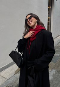 Čierny nadrozmerný kabát cez čierny sveter v kombinácii s červeným šálom. Oblečenie dopĺňajú čierne slnečné okuliare a malá čierna kabelka.