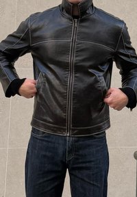 Άνδρας που φοράει μαύρο δερμάτινο μπουφάν με φερμουάρ και σκούρα τζιν, με τα χέρια στις τσέπες, στέκεται μπροστά σε ένα πλ ακιδωτό ανοιχτό γκρι τοίχο.