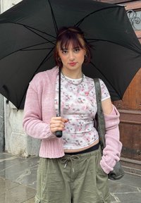 Νεαρό άτομο που κρατά μαύρη ομπρέλα φοράει ροζ πουκάμισο με σχέδια γάτες, ροζ πλεκτό πουλόβερ, χακί παντελόνια και κουβαλάει γκρι τσάντα ώμου.