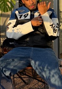 Melnā un baltā ādas jaka ar zilām detaļām, dekorēta ar numuru 19 un sānu uzlīmēm. Zilas džinsa bikses. Sēž uz krēsla dabiskā apgaismojumā.