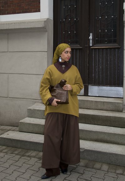 Mujer con un pañuelo amarillo en la cabeza y un conjunto amplio de tonos mostaza y marrón sostiene un bolso de cuero marrón, de pie sobre escalones de concreto junto a una puerta de madera oscura.