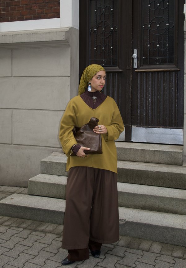 Frau mit gelbem Kopftuch und locker sitzender, senfgelber und brauner Kleidung hält eine braune Ledertasche und steht auf Betontreppen vor einer dunklen Holztür.
