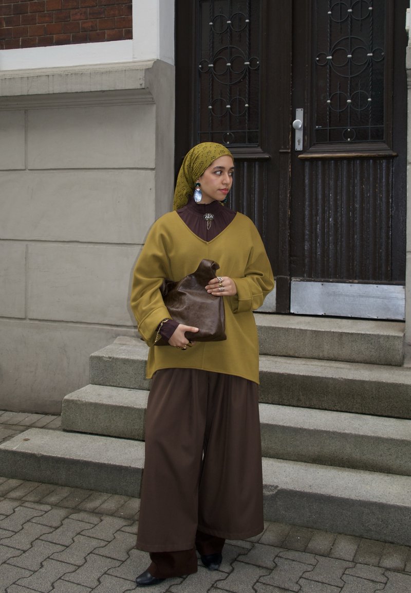 Femme portant un foulard jaune et une tenue ample moutarde et marron, tenant un sac en cuir marron, debout sur des marches en béton près d'une porte en bois sombre.