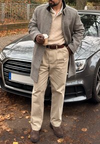 Grå rutete kåpe over en kremfarget kabelstrikket genser, beige bukser, brune hansker og brune støvler, holder en kaffekopp i en utendørs setting.