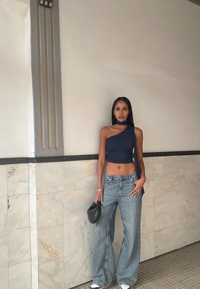 Top corto de un solo hombro en color navy combinado con jeans de mezclilla claros de pierna ancha. La modelo sostiene un bolso texturizado negro y está de pie frente a una pared de mármol.