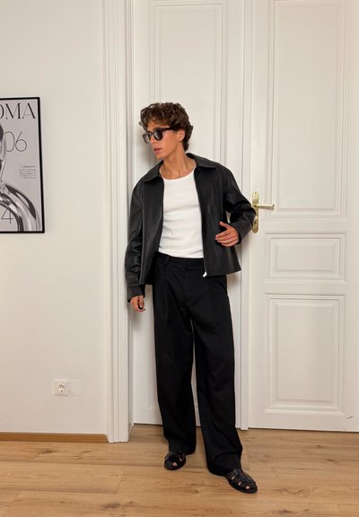 Chaqueta de cuero negra con cremallera sobre una camiseta blanca, combinada con pantalones negros holgados y sandalias negras. Cabello rizado y gafas de sol oscuras.