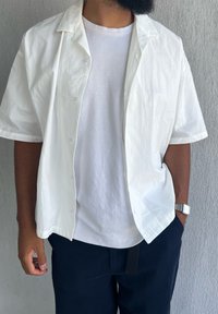 Chemise blanche à manches courtes avec une coupe décontractée, design à boutons, et texture naturelle, portée sur un t-shirt blanc uni et un pantalon foncé.