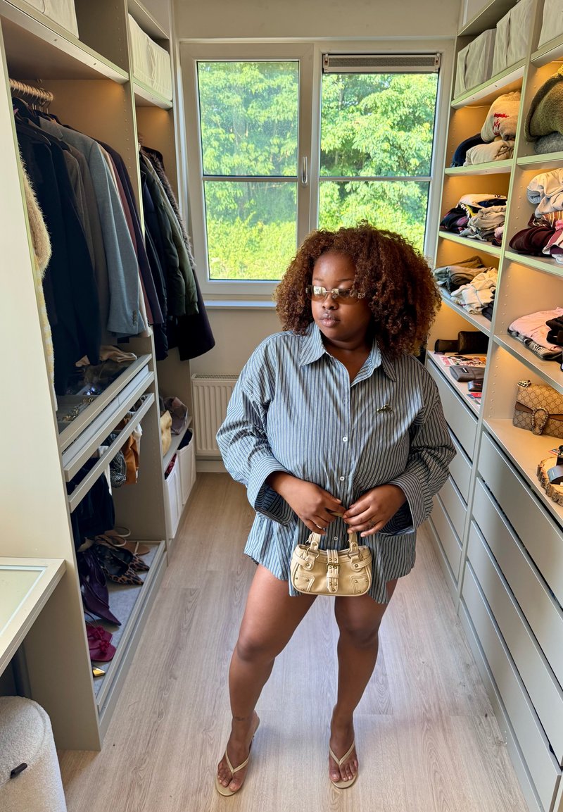 Femme aux cheveux bouclés et lunettes, portant une chemise rayée surdimensionnée, tenant un petit sac à main beige dans un dressing avec des vêtements et des étagères.