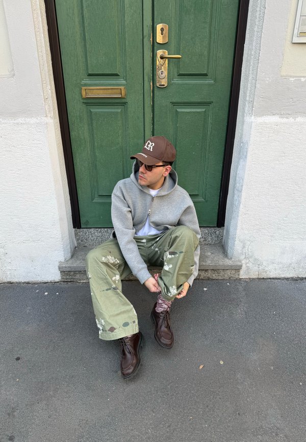 Homme portant un sweat à capuche gris, un pantalon éclaboussé de peinture verte, des chaussures marron et une casquette marron, assis sur le seuil d'une porte, ajustant sa chaussette.