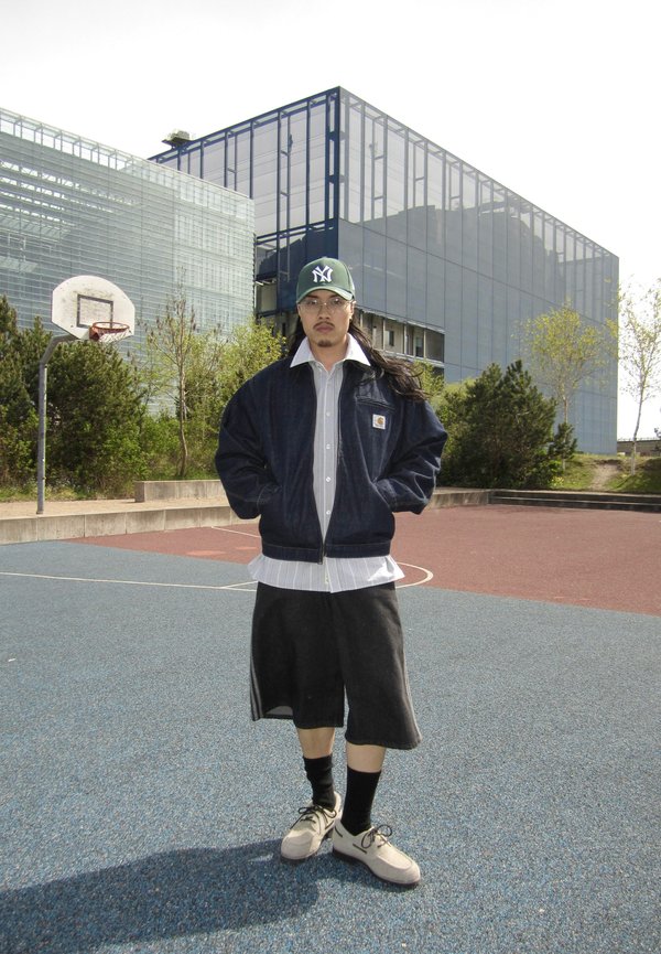 Mann mit Baseballkappe, Brille, Jacke und Shorts steht auf einem Outdoor-Basketballplatz mit modernen Glasgebäuden im Hintergrund.