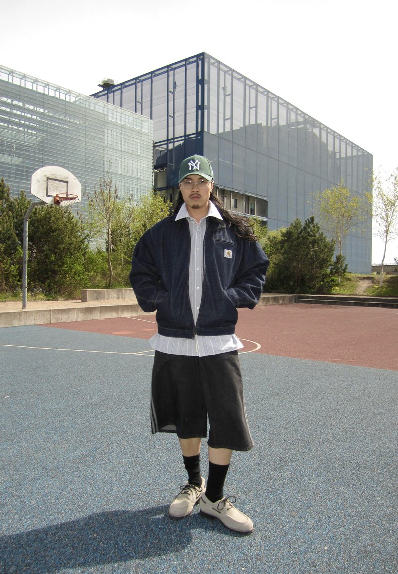 Mand iført en kasket, briller, jakke og shorts står på en udendørs basketballbane med moderne glasbygninger i baggrunden.