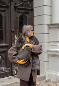 Barna gyapjúkabát szőrme gallérral, mustár színű pulóver, barna kesztyűk és szürke táska. A háttérben díszes faajtó és világosszürke fal látható.