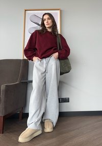 Bourgogne oversized sweatshirt, grå joggingbukser, beige tøfler. Modellen poserer med en olivengrøn taske foran en minimalistisk baggrund.