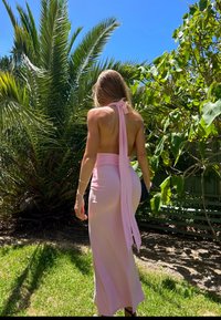 Une femme en robe rose dos nu à col halter se tient sur l'herbe près de palmiers et d'arbres feuillus sous un ciel bleu clair.