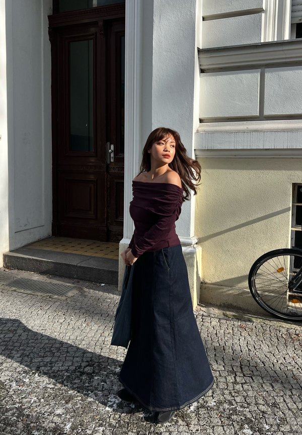 Mujer con largo cabello oscuro y blusa morada con los hombros descubiertos, de pie en una calle empedrada junto a un edificio blanco y una puerta de madera, con la luz del sol proyectando sombras.