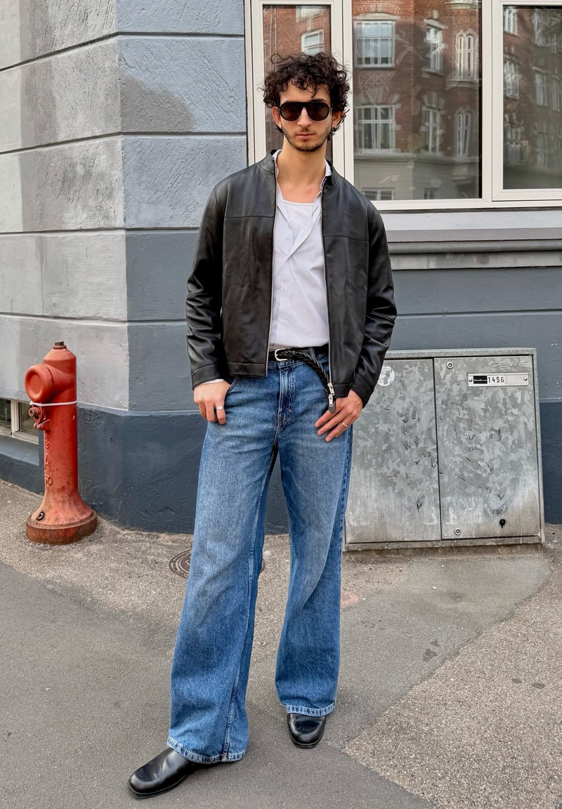 Casaco bomber de couro preto, camisa de riscas brancas, jeans de denim azul largo, botas pretas, óculos de sol. Cenário urbano com um hidrante vermelho.